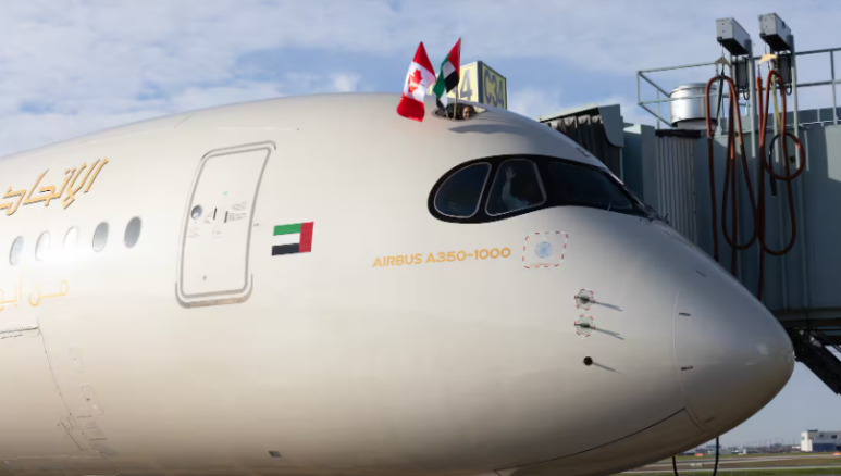 Etihad A350 at a gate with crew member holding Canadian and UAE flags from open door above cockpit.