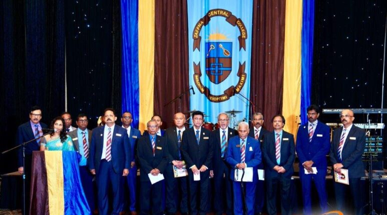 Group of alumni stand in front of their Jaffna Central College flag on stage.