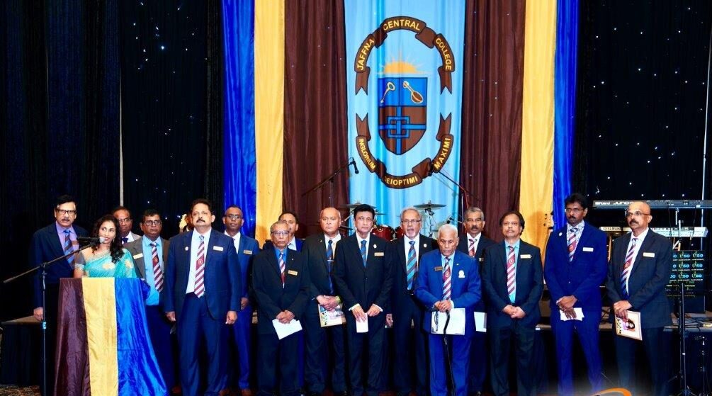 Group of alumni stand in front of their Jaffna Central College flag on stage.